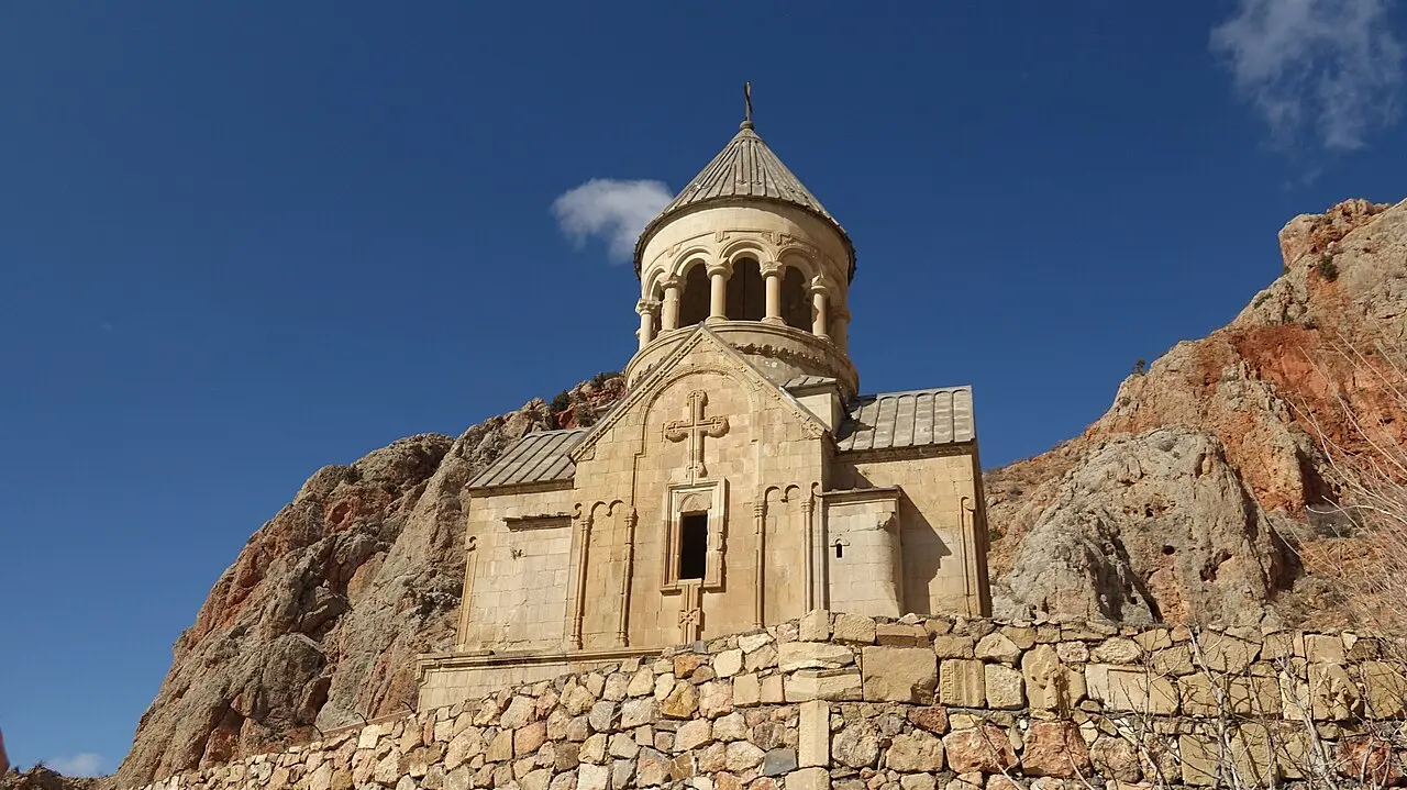 A domed medieval church at Noravank monastery standing against a clear blue sky, with red-brown rock rising behind