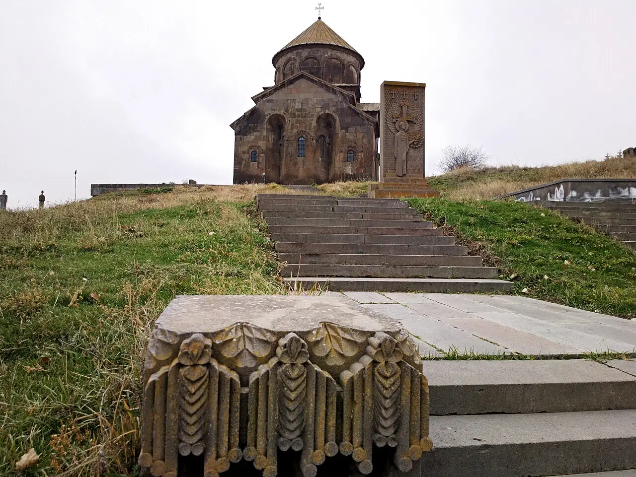 The domed 7th-century Sisavan church above Sisian, with a carved khachkar and old stone fragments on the grass in front