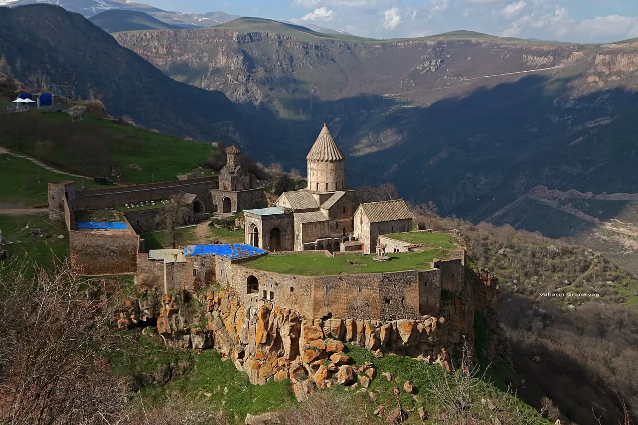 Tatev monastery on its basalt plateau at the edge of the deep Vorotan gorge, mountains rising behind under a bright sky