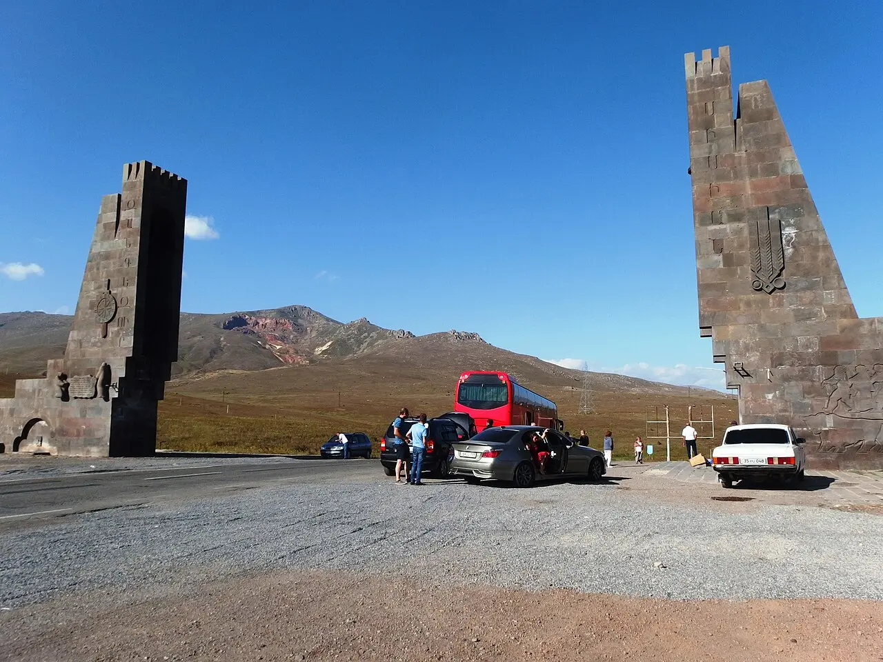 The two tall carved stone pylons of the monument on the Vorotan Pass summit, with cars and a bus stopped on the bare highland