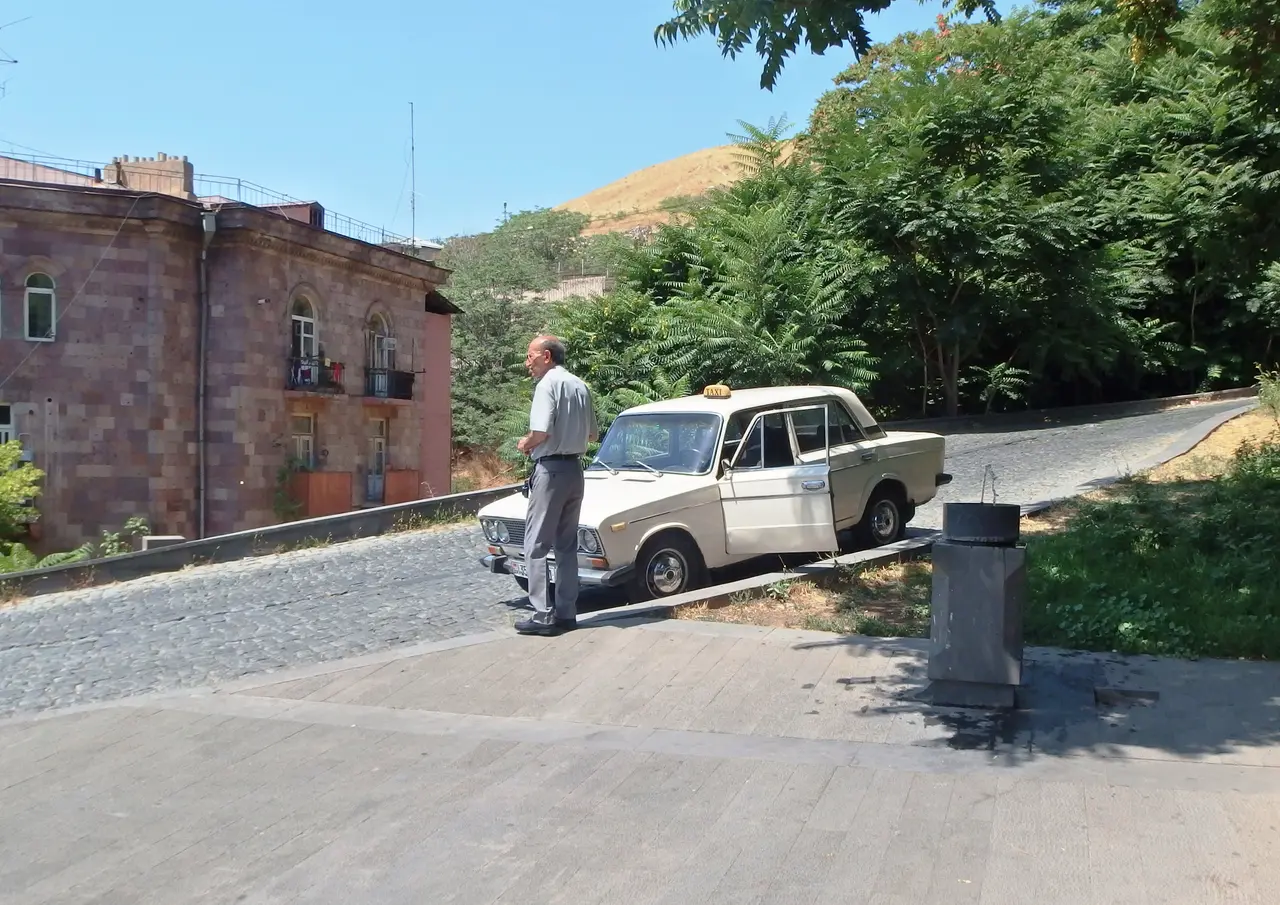 An old white Soviet-era Lada taxi with a driver standing beside it on a Yerevan street