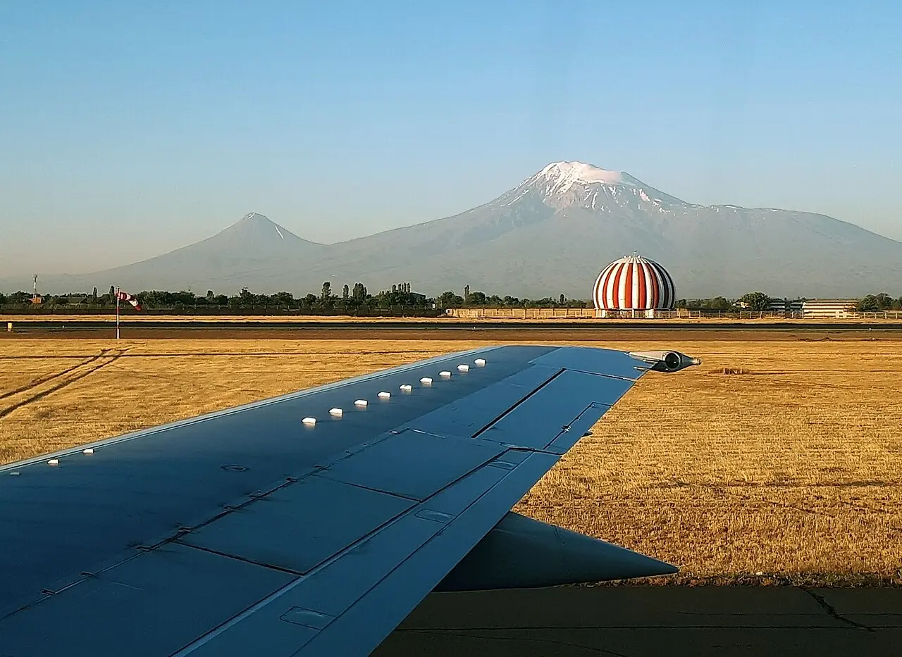 The wing of a departing plane at Zvartnots airport with Mount Ararat and the round airport building behind on a clear morning