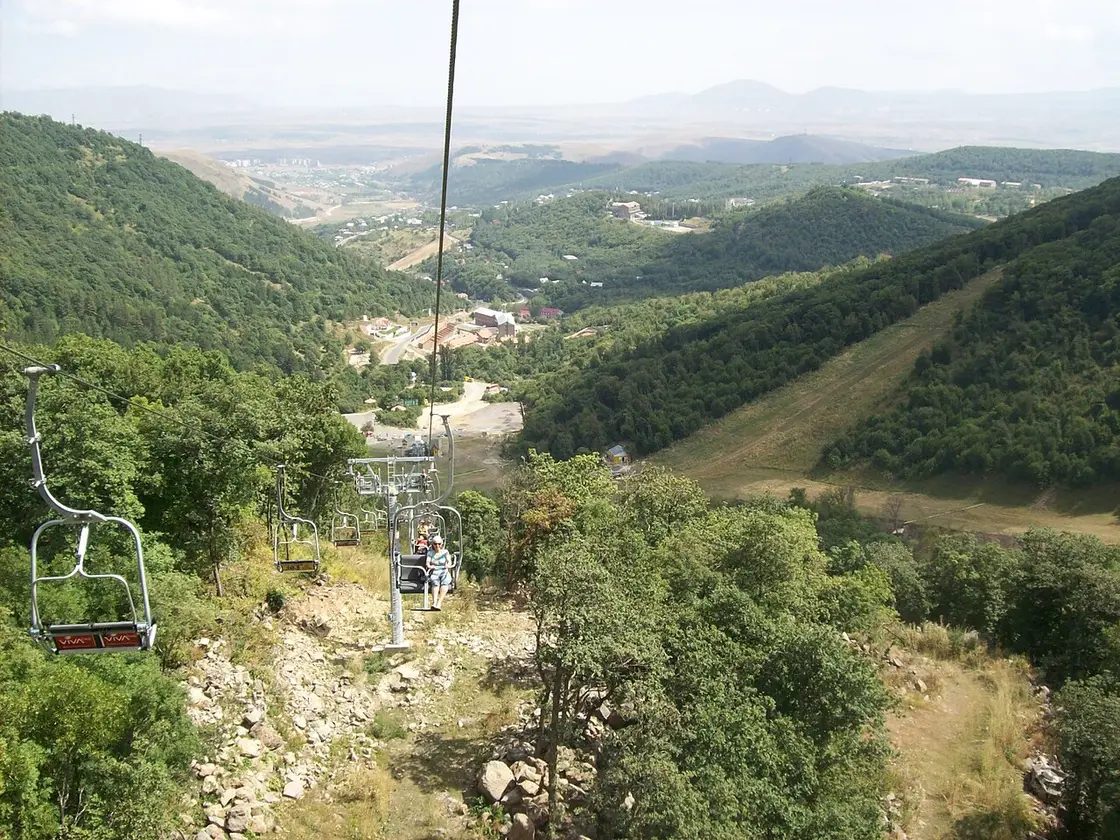 The same Tsaghkadzor chairlift in summer over green forested slopes with the valley and town in the distance