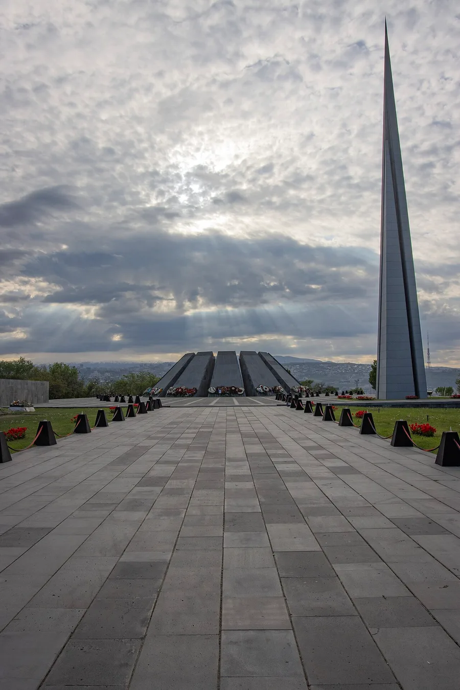 Wreaths and flowers piled at the base of the twelve slabs on the Tsitsernakaberd plaza, the stele and red tulips beyond, under a dramatic sky