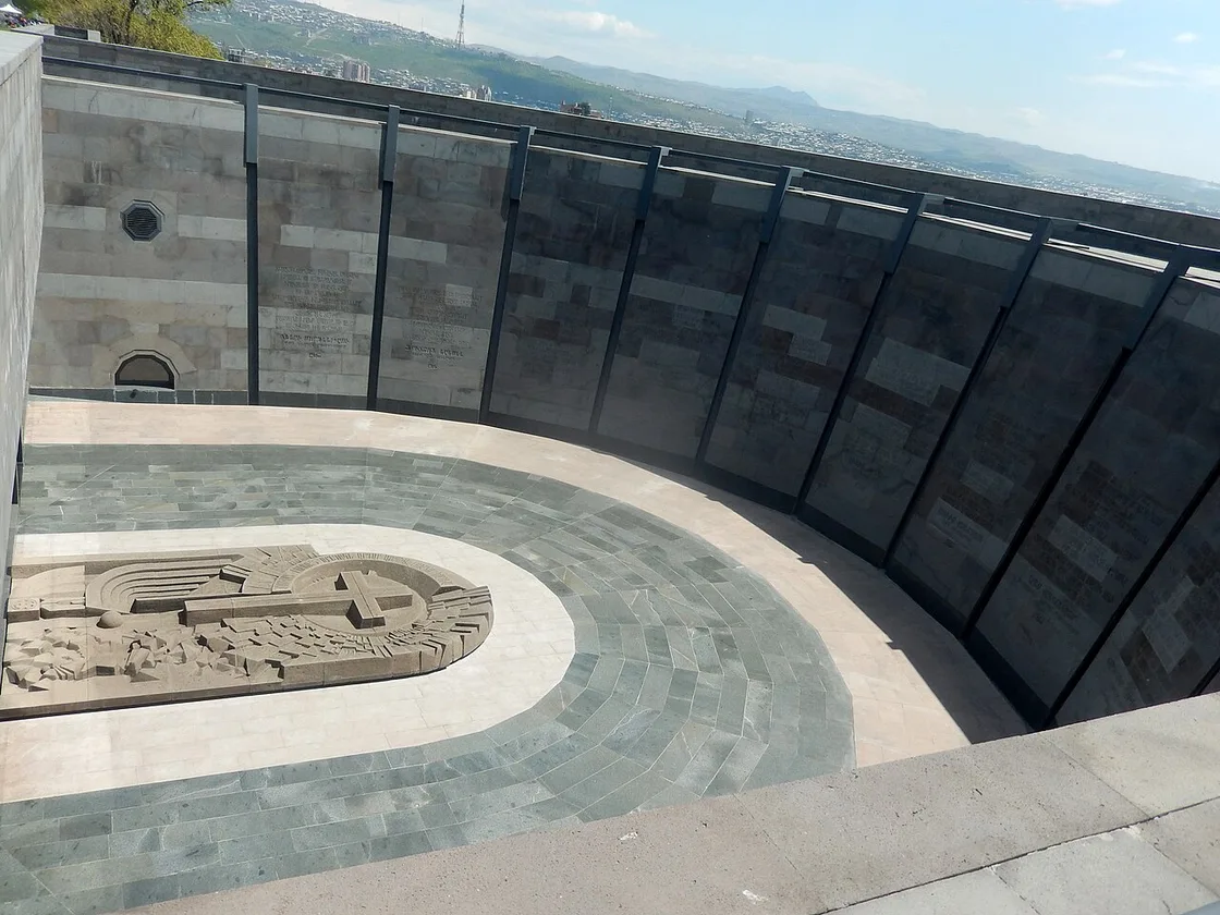 A curved stone terrace at Tsitsernakaberd with an inscribed wall and a carved relief, looking out over Yerevan and the hills