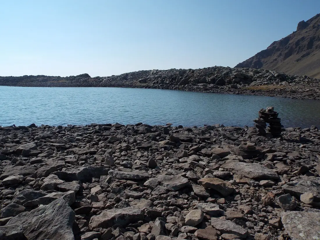 Dark broken lava along the shore of Ughtasar lake with a small stone cairn and a steep rocky ridge beyond