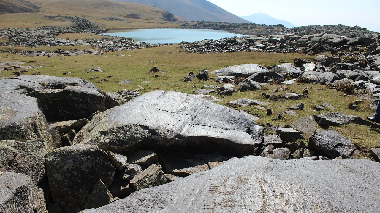 Carved volcanic boulders in the foreground and the small crater lake of Ughtasar in a treeless high basin under a blue sky