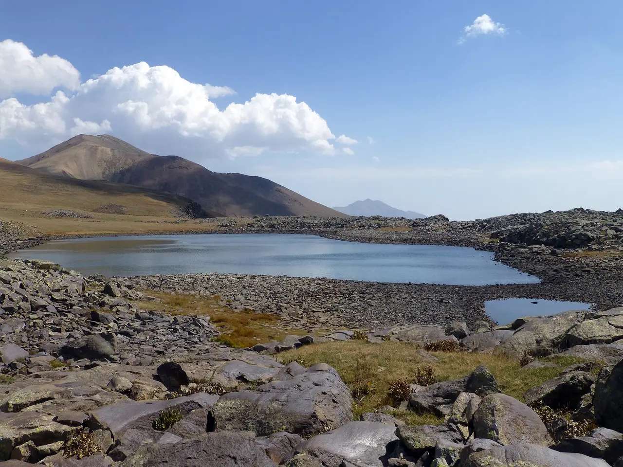 The small high crater lake at Ughtasar ringed by dark volcanic boulders and treeless brown ridges under a bright sky