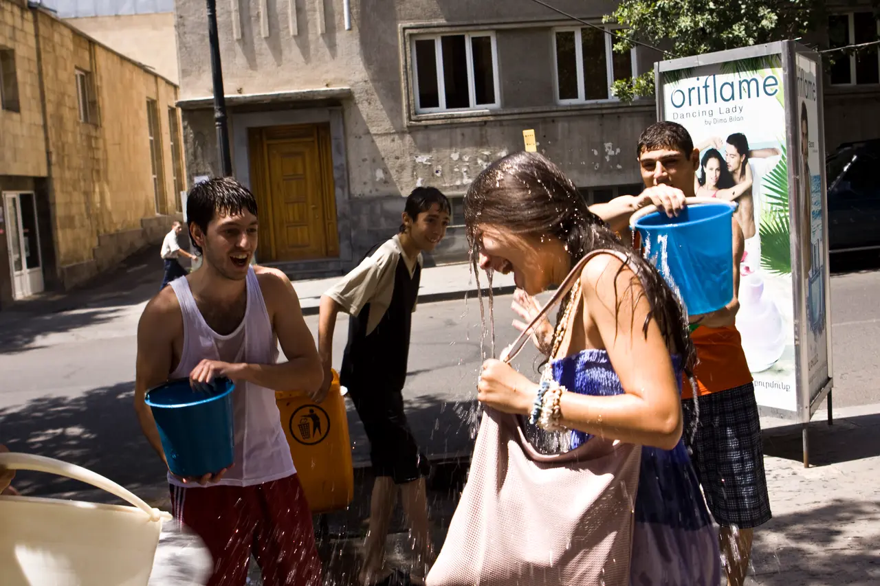 A group of young people with buckets soaking a woman with a shoulder bag on a Yerevan sidewalk during Vardavar