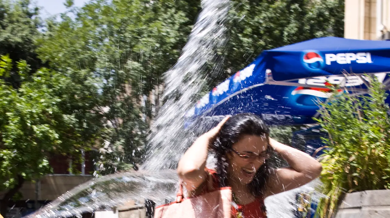 A woman in a red dress on a Yerevan street being hit at once by a bucket and a hose of water during Vardavar