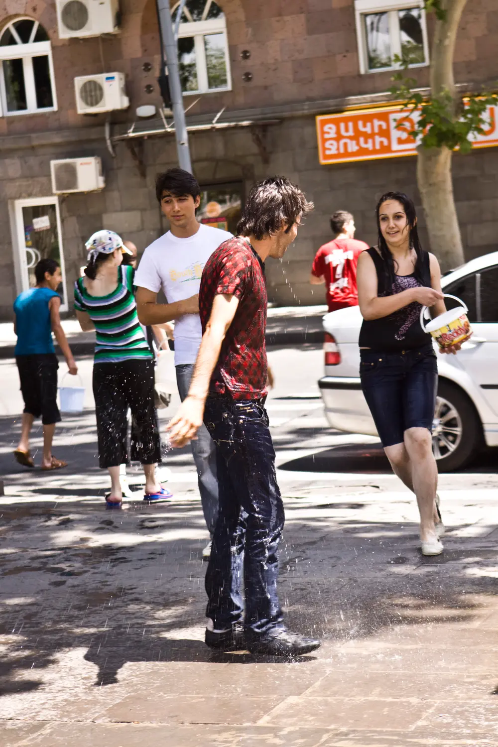 A soaked man standing at a Yerevan street corner as people carry buckets of water past him on Vardavar