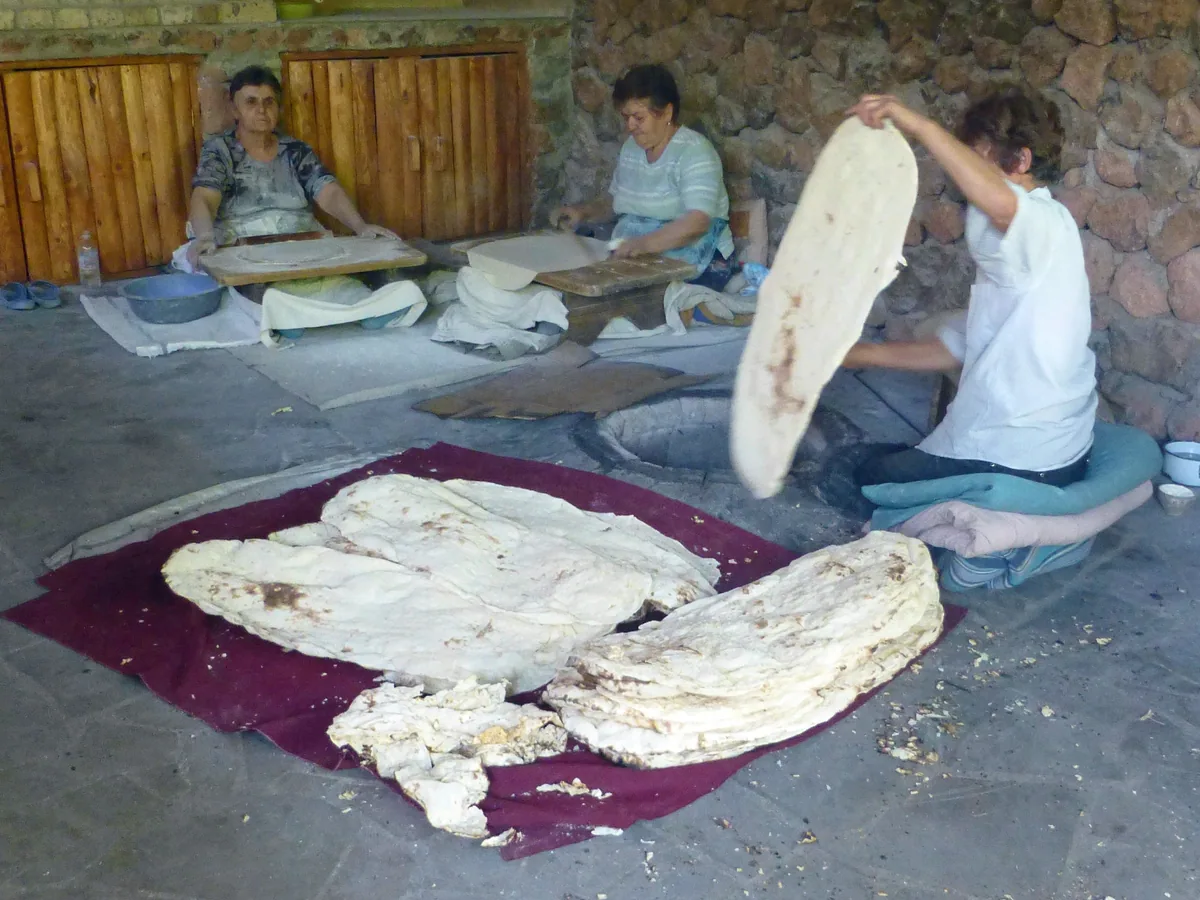 Three women baking lavash flatbread by hand at a village tonir oven, with baked sheets stacked on a red cloth