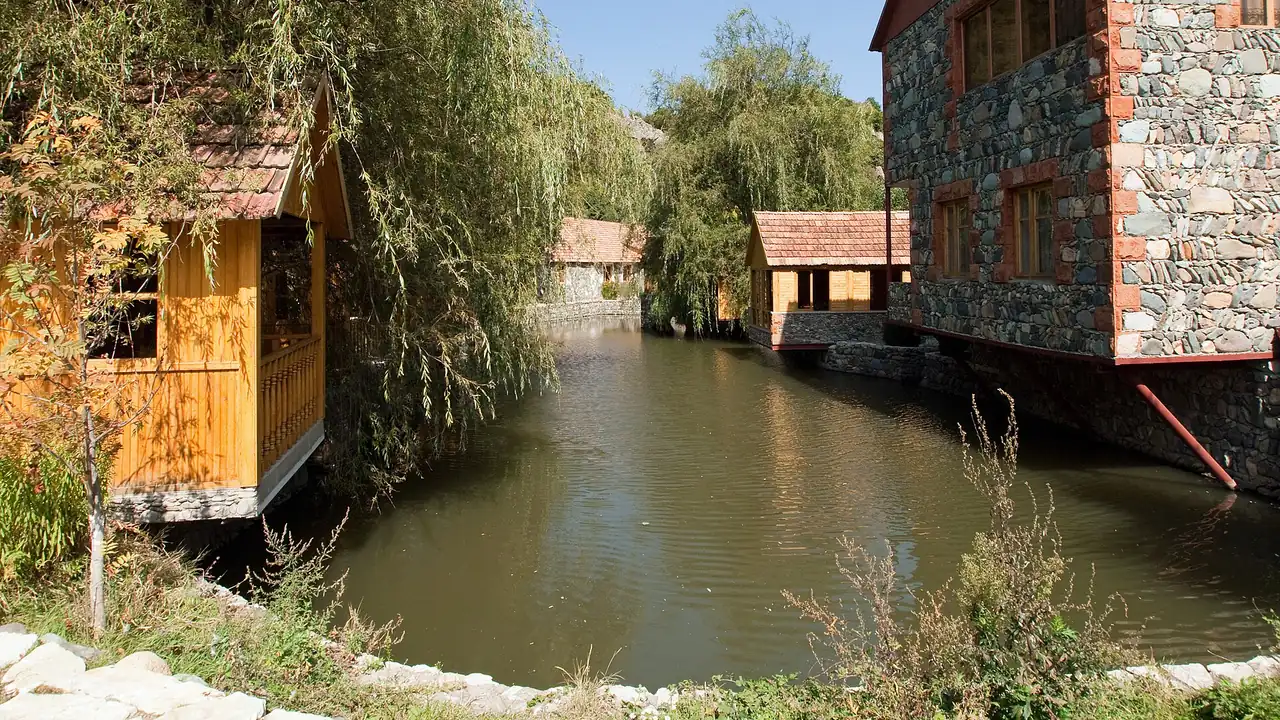 Wooden and stone guesthouses standing over a still pond ringed by willow trees in Dilijan, Armenia