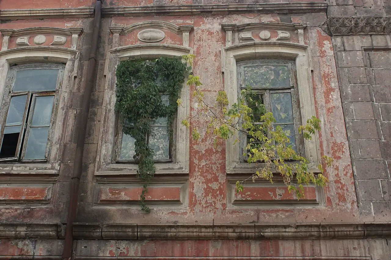 A peeling red-tuff facade in Gyumri with carved window surrounds and ivy growing over the glass