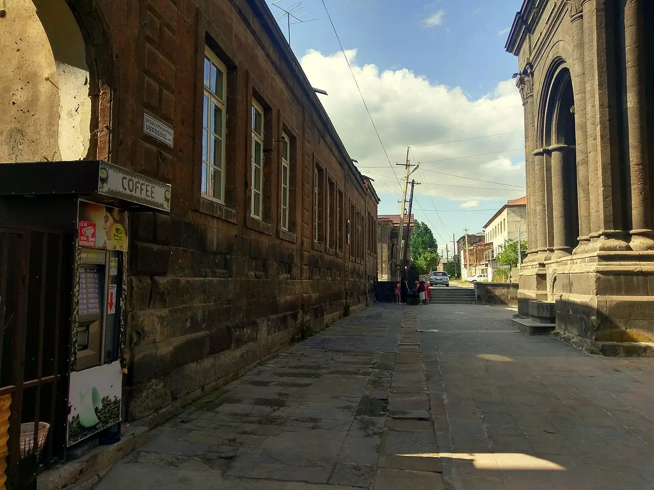A paved old-town street in Gyumri with tuff buildings, a coffee kiosk and a church under a blue sky