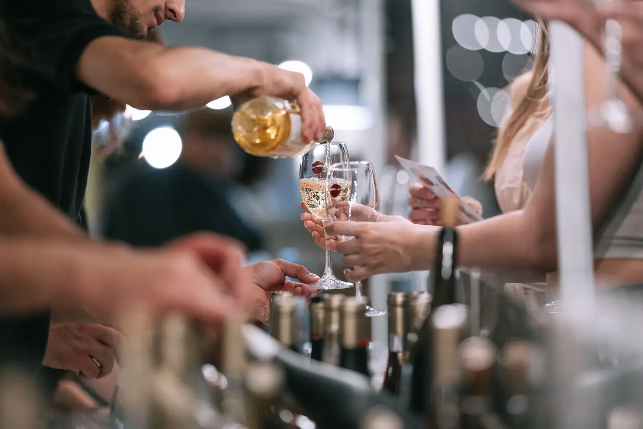 A close-up of white wine being poured into a glass at a tasting table lined with bottles