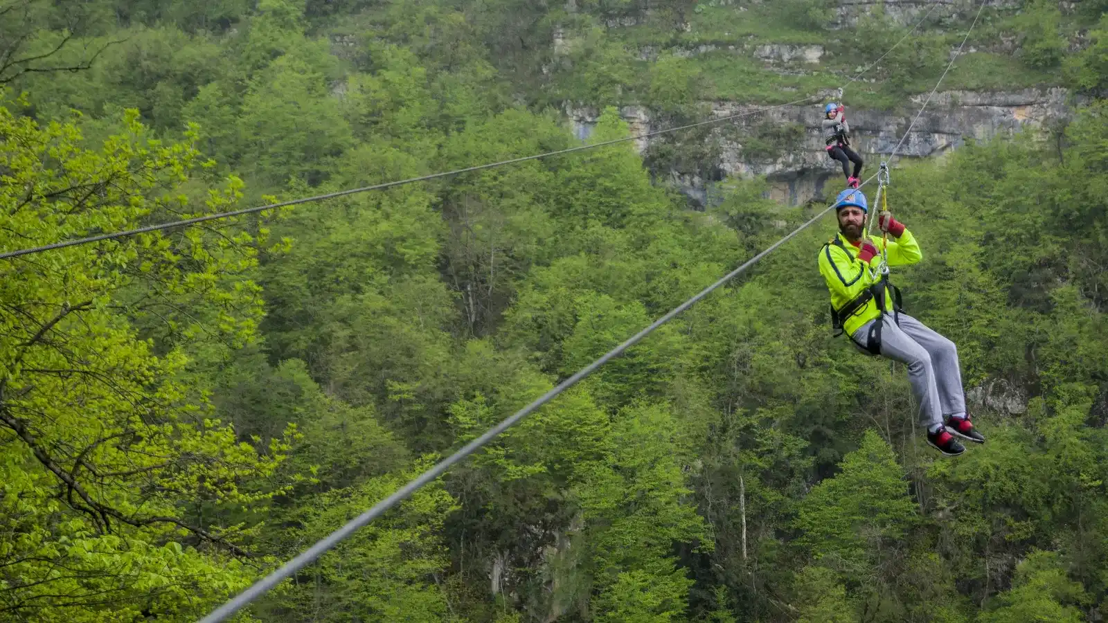 Two riders in helmets and harnesses hanging from a zipline cable above the green forested Yenokavan canyon