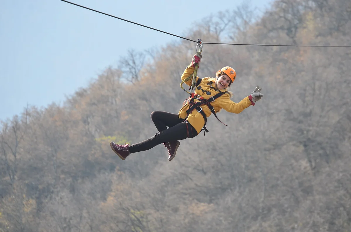 A smiling woman in a yellow jacket and orange helmet riding the zipline cable above bare autumn trees