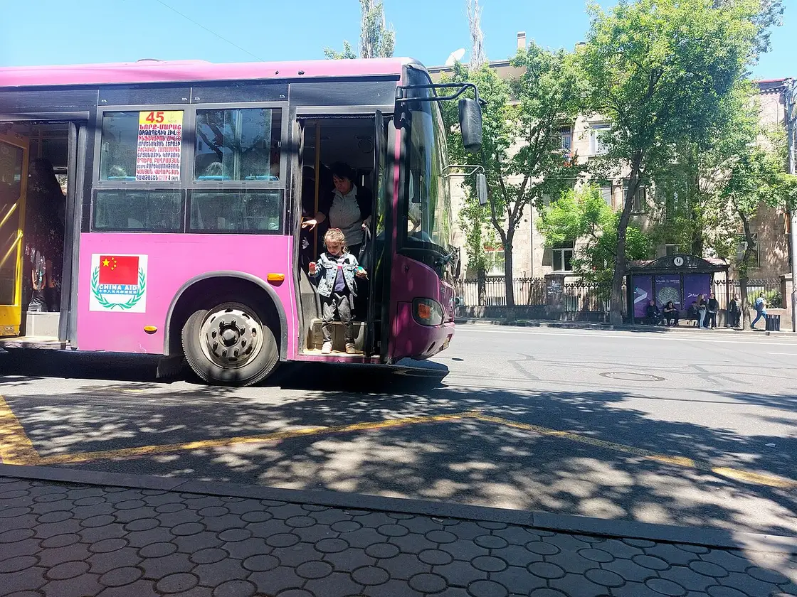 A pink Yerevan city bus stopped at a leafy street stop with a passenger stepping down from the front door