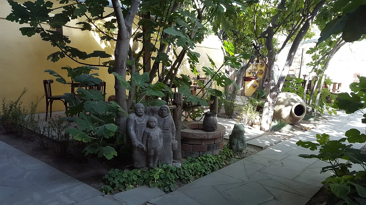 A courtyard art cafe in Yerevan shaded by fig trees, with stone folk-art sculptures and a large clay karas jar among the greenery