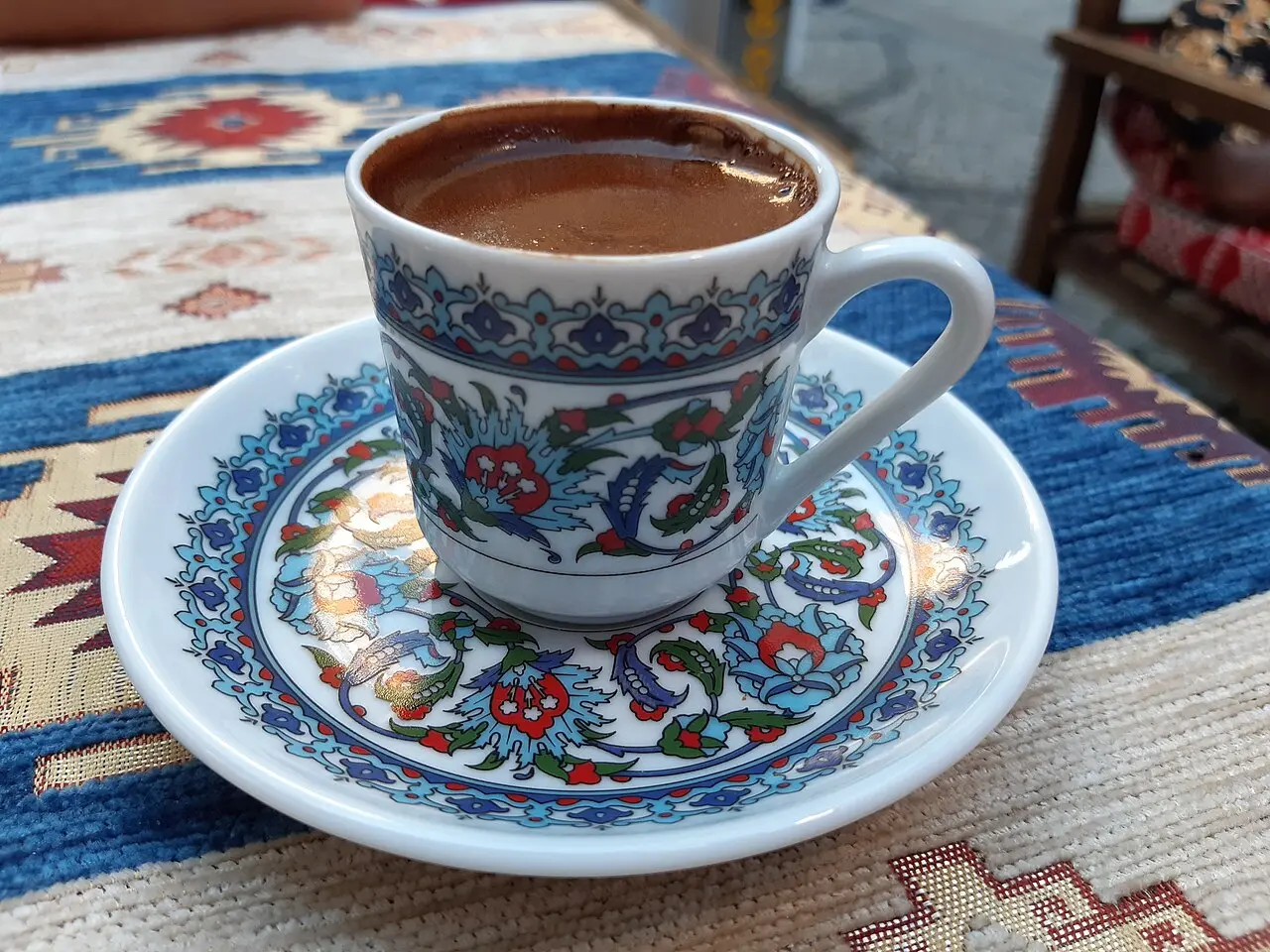 A porcelain cup of strong dark coffee with foam on top, served on a matching floral saucer over a patterned kilim rug