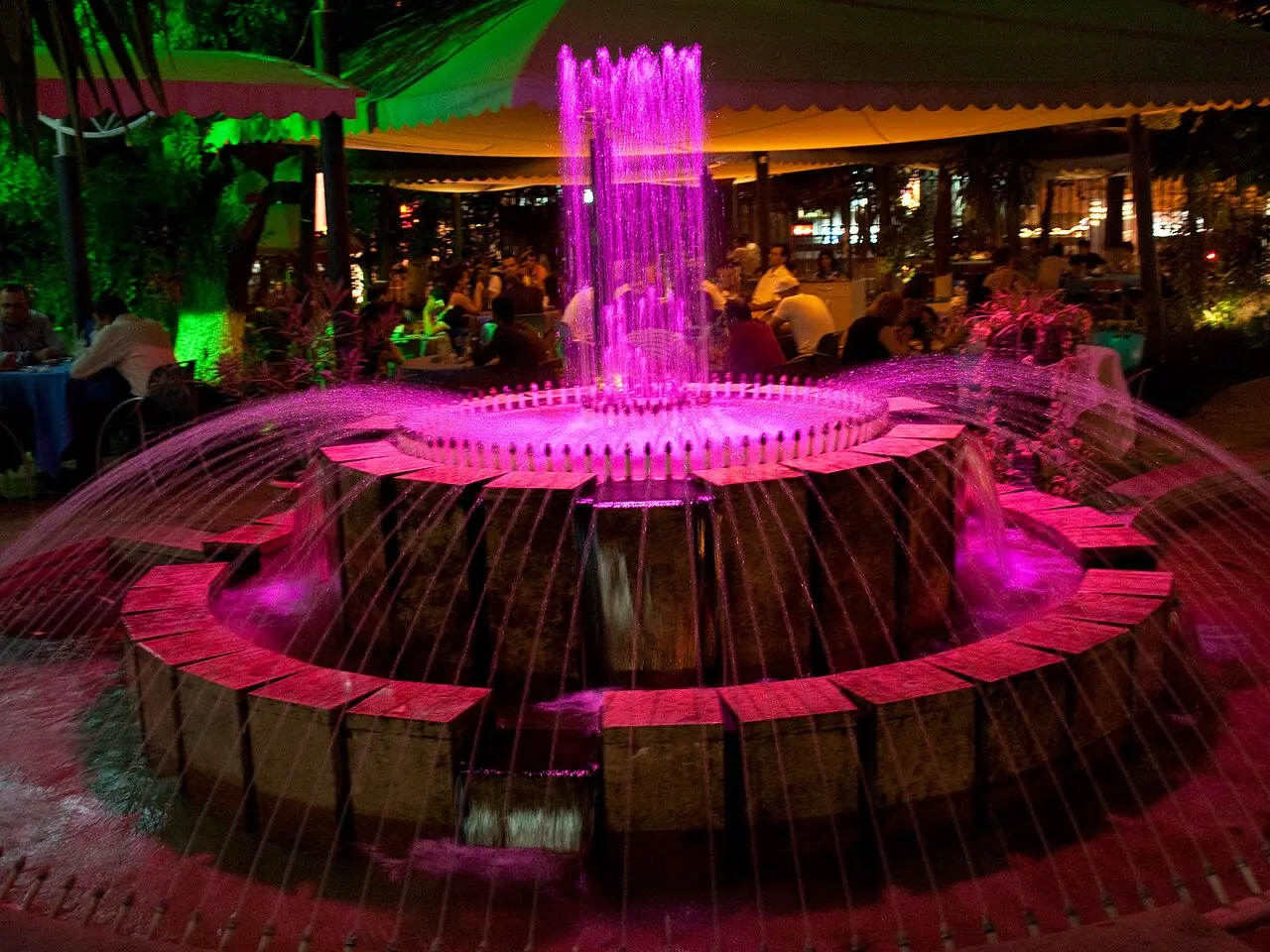 A colour-changing fountain lit magenta at night in a Yerevan cafe garden, with diners seated at tables under the awning behind it