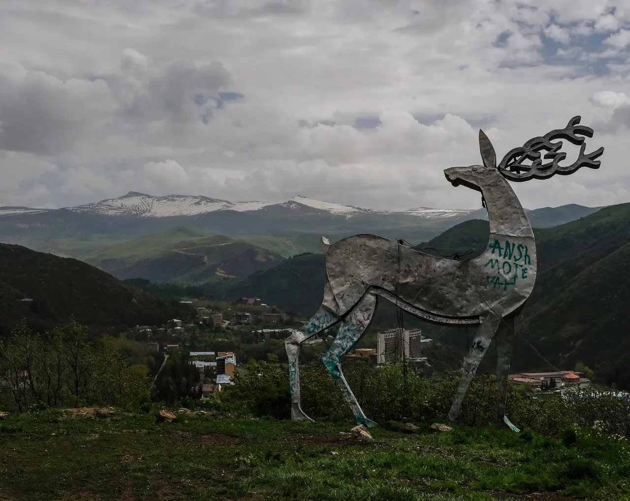 A metal deer statue on a hillside above Jermuk with the spa town and snow-streaked mountains behind